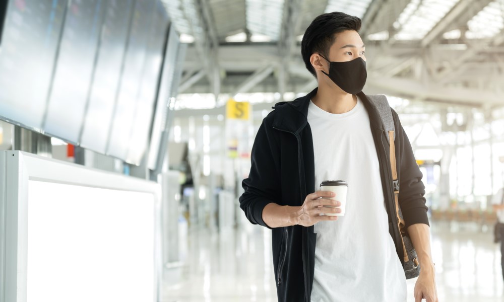 A casually dressed young man in a black cloth mask stands in a sunny airport terminal.