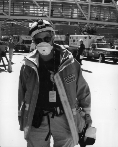A Boston EMS physician in a hard hat, mask, and heavy jacket stands in a parking lot with a row of ambulances in the background.