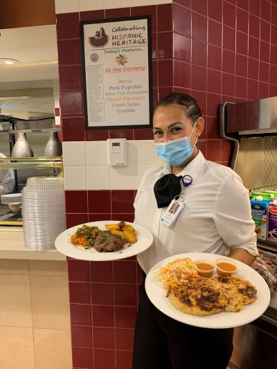Flor Chavez holding plates of food in front of a Celebrating Hispanic Heritage sign at a Mass General cafeteria.
