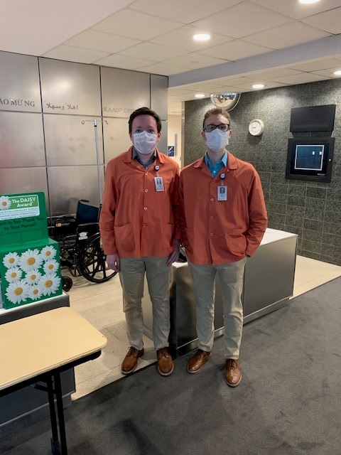 Two men in volunteer jackets and face masks stand by the information desk in a hospital lobby.