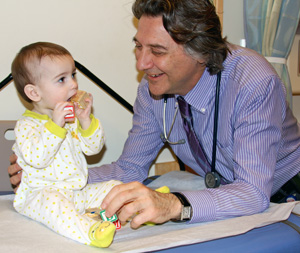 A doctor smiles at a onesie-clad baby who's sitting up on an examination table.
