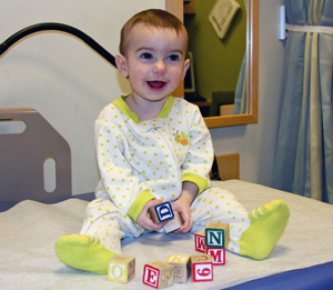 A beaming baby in a onesie sits on an examining table and plays with blocks.