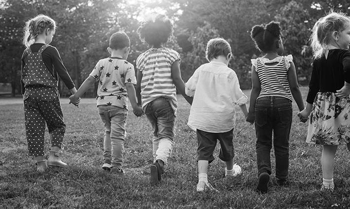 Photo of six school-aged children in a field holding hands and walking away from the camera