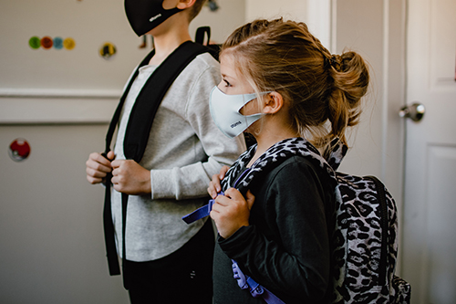 Two children are ready to go to school as they wear masks and backpacks.