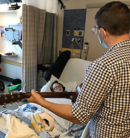 Adam Sankowski, a music therapist at MGfC, stands at Molly Murphy's bedside at MGfC strumming a guitar while Molly, who is in her bed, sings along. 