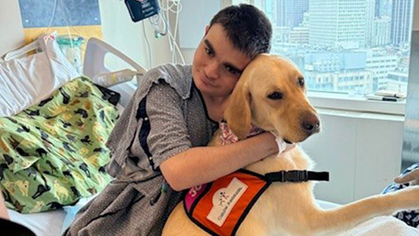 Pediatric patient Matthew hugs Kayce the facility dog while sitting on a hospital bed.