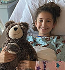 11-year-old Lindsey smiles and hugs a teddy bear in her hospital bed.