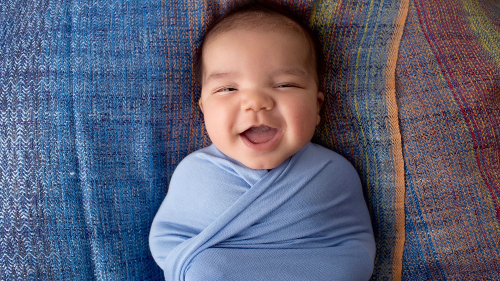 A smiling baby swaddled in blue lies on a many-colored blanket