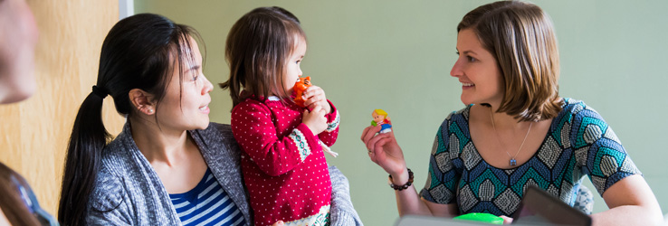 A toddler standing on her mother's lap holds a toy to her mouth while considering a different toy offered to her by Dr. Lauren Fiechtner.