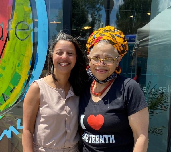 Deb Washington, an older Black woman wearing a T-shirt that says 'I heart Juneteenth,' stands in front of a cafe with a smiling younger woman.