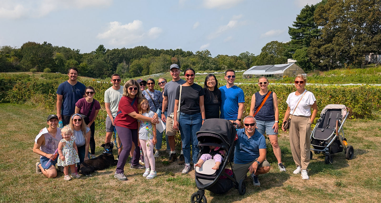 A group pf happy people (plus a child and two toddlers) pose beside a hedge at a farm.