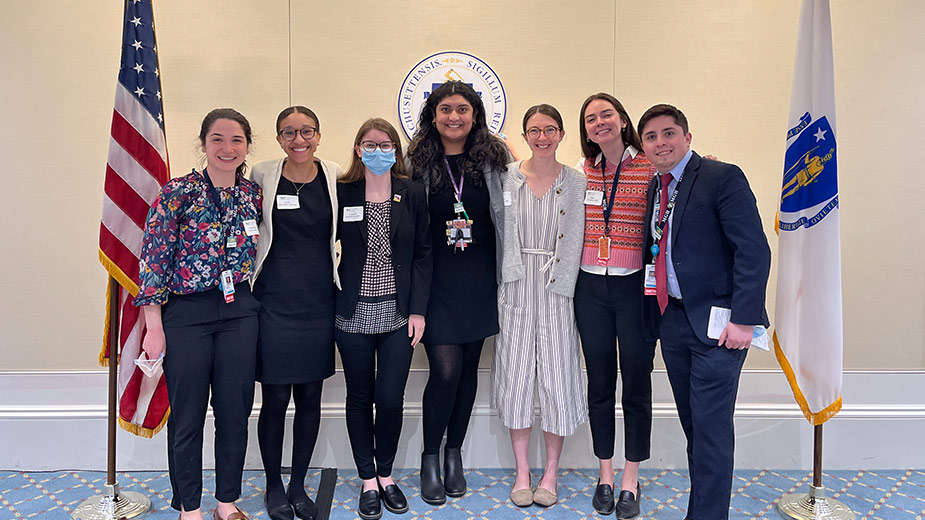 7 hospital residents standing together for a group photo at the Massachusetts state house with an American flag on the left and the Massachusetts state flag on the right