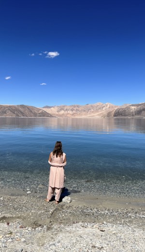 Dressed in a white salwar khameez, Dr. Singh stands at the edge of a lake. The blue skies and desert mountains in the distance are reflected in the lake.