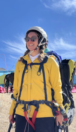 A grinning Dr. Singh in a yellow jacket and white helmet, wearing a paragliding harness, with the paraglider strapped to her back.