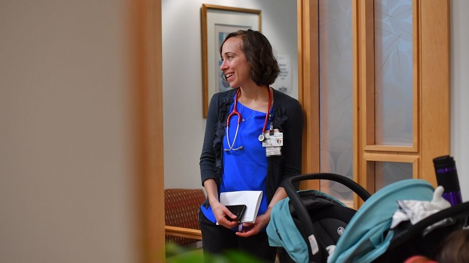 A doctor, with a stethoscope around her neck, smiles while looking back through a doorway. 