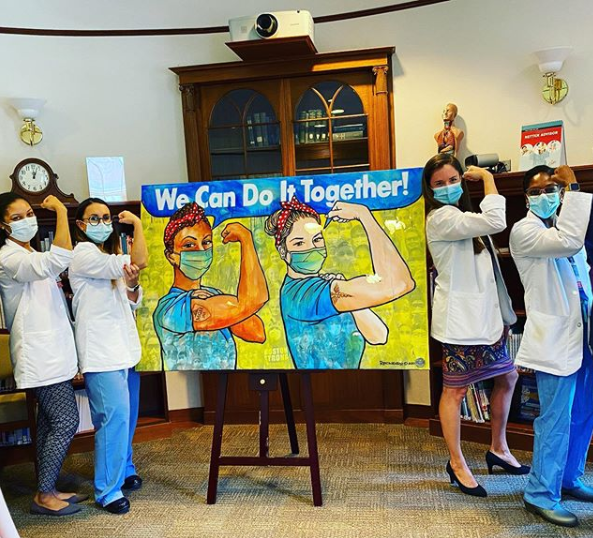 A group of four female surgical residents pose with artwork that is dedicated to the incredible work of Mass General staff during COVID-19.