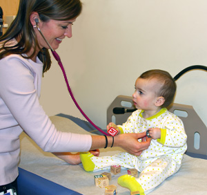 A onesie-clad baby with a block in one hand fingers the stethoscope as a smiling doctor presses the stethoscope to its chest.