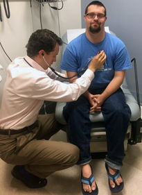 A young man with Down syndrome sits on an examination table, looking faintly, patiently amused, while a researcher listens to his heart with a stethoscope.