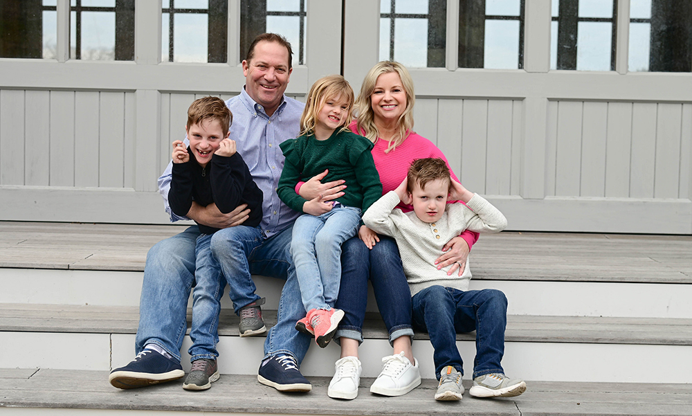The Hartford family sits on a wooden deck, smiling for the camera. The Hartford family sits on a wooden deck, smiling for the camera.