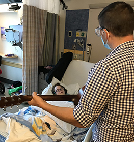 Adam Sankowski, a music therapist at MGfC, stands at Molly Murphy's bedside at MGfC strumming a guitar while Molly, who is in her bed, sings along.