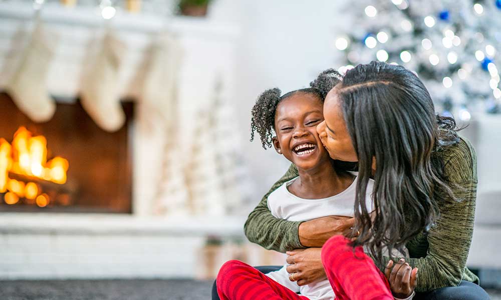 A smiling child is embraced by her mother with Christmas decorations in the background A smiling child is embraced by her mother with Christmas decorations in the background
