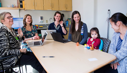 A toddler, her mother, and four smiling members of her treatment team sit around a table in an exam room.