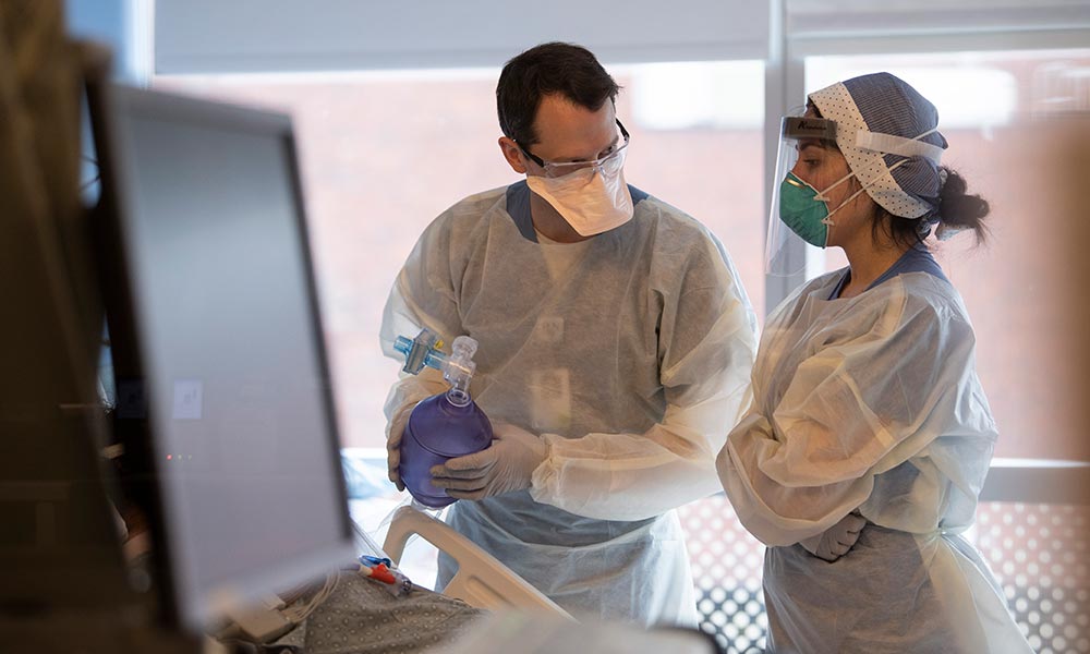 Two gowned and masked medical staff confer at a patient's bedside. Two gowned and masked medical staff confer at a patient's bedside.