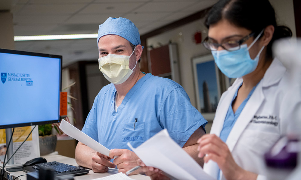 Two medical professionals: One smiling at the camera from behind a mask, the other is studying a patient chart and wearing a mask. Two medical professionals: One smiling at the camera from behind a mask, the other is studying a patient chart and wearing a mask.