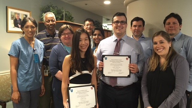 A young man and a young woman holding up their diplomas, with part of the department posing behind them. A young man and a young woman holding up their diplomas, with part of the department posing behind them.