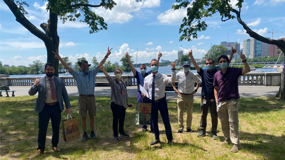 In a Boston park with a waterway in the background, a man holds his diploma in one arm and gives a thumbs-up with the other hand. Around him, other people from the department give thumbs-ups. In a Boston park with a waterway in the background, a man holds his diploma in one arm and gives a thumbs-up with the other hand. Around him, other people from the department give thumbs-ups.