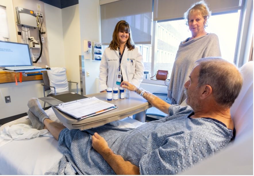 A patient sits in bed with medications while two women look on A patient sits in bed with medications while two women look on