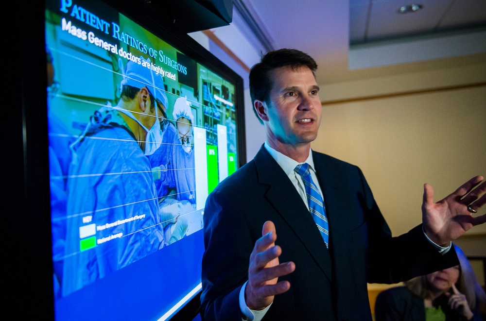Matthew Hutter makes a presentation in front of a screen displaying a chart and a photo of surgeons in surgery Matthew Hutter makes a presentation in front of a screen displaying a chart and a photo of surgeons in surgery