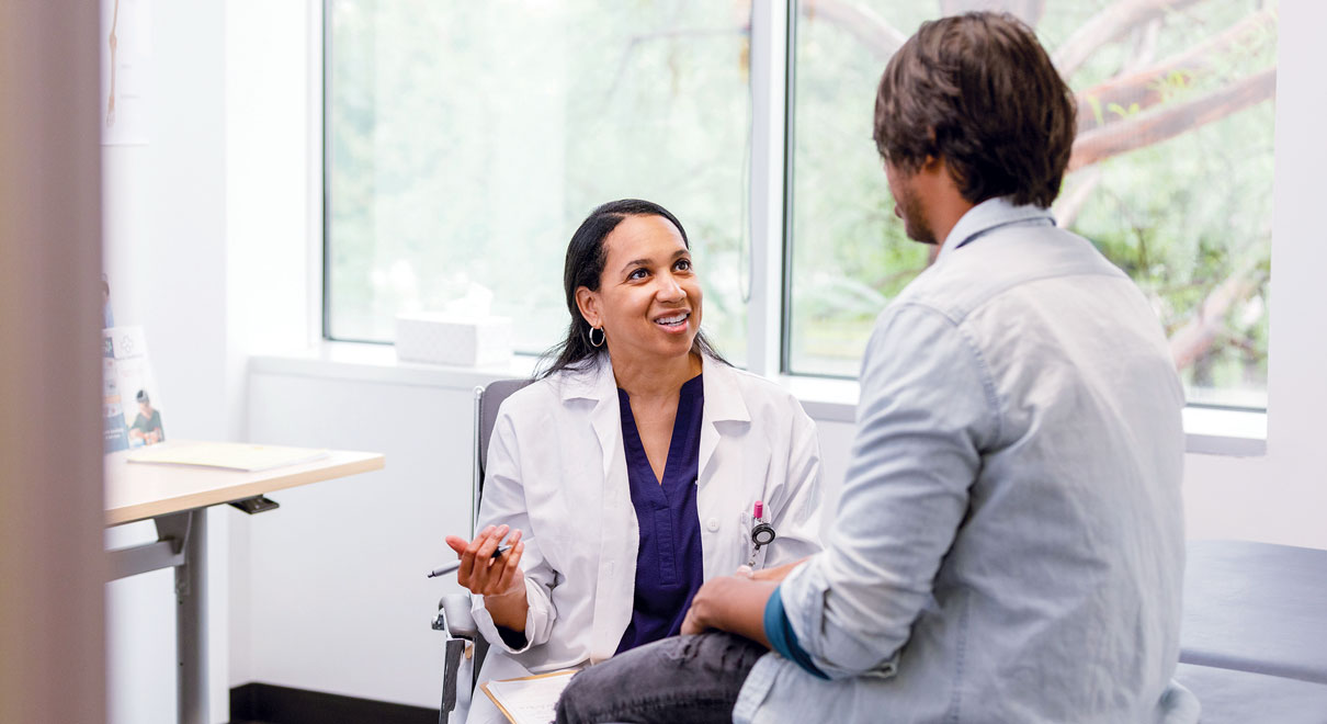 A patient sits on an exam table, talking with a provider seated across from him A patient sits on an exam table, talking with a provider seated across from him